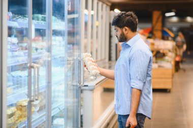 Çeşitli ürünlerle çevrili bir marketin soğutucu koridorunda alışveriş yaparken taze yumurta seçen odaklanmış bir müşteri.