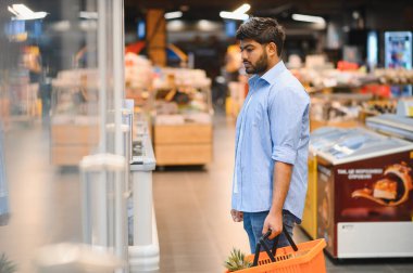 Market alışverişi yapan genç Hintli bir adam, elinde bir sepet ve dondurulmuş gıda ürünlerini süpermarket buzdolabı bölümünden seçiyor.