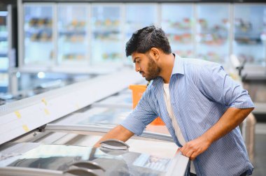 Donmuş yiyecek seçen konsantre olmuş genç Hintli bir adam süpermarkette bir dondurucunun cam kapısını açıyor.