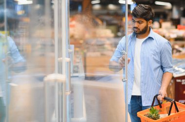 Odaklanmış Hintli müşteri buzdolabı kapısını açıyor, alışveriş sepetini marketten taze ürünleri seçiyor.