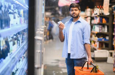Elinde alışveriş sepeti olan Hintli bir müşteri süpermarket buzdolabından ürün seçiyor.