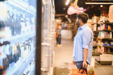 Market alışverişi yapan genç Hintli bir adam, elinde bir sepet ve süpermarket buzdolabı bölümündeki ürünlere bakıyor.