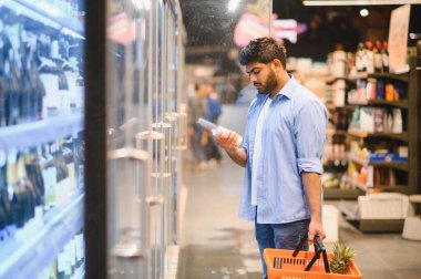 Genç Hintli bir adam süpermarket buzdolabı koridorunda alışveriş yaparken bir şişeyle ilgili bilgileri dikkatle okuyor.