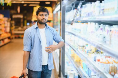 Genç Hintli adam alışveriş arabasını itiyor ve süpermarketin dondurulmuş bölümünde süt ürünlerini seçiyor.