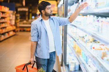 Genç Hintli adam alışveriş sepetini tutuyor ve marketin dondurucu bölümünden süt ürünlerini seçiyor.