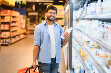 Bakkal alışverişi yapan genç Hintli adam, süpermarket süt ürünleri bölümünde süt şişesi ve sepeti tutuyor.