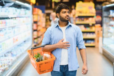 Genç Hintli bir adam bir alışveriş sepeti taşıyor, dolu bir marketin canlı koridorlarında geziniyor.