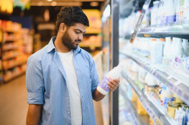 Elinde süt şişesi olan odaklanmış bir müşteri marketin dondurulmuş bölümündeki farklı süt markalarını karşılaştırıyor.
