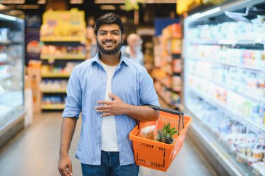 Genç Hintli bir adam bir alışveriş sepeti taşıyor, market koridorunda geziniyor, taze ürünler ve sağlıklı malzemeler seçiyor.