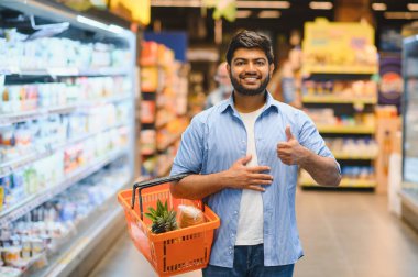 Süpermarkette alışveriş sepeti taşırken mutlu müşteri başparmak hareketi yapıyor.