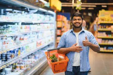 Mutluluk dolu bir alışveriş sepetini süpermarkette tutarken başparmağını kaldıran mutlu müşteri.