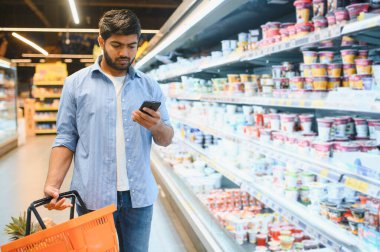 Genç Hintli bir adam akıllı telefon kullanıyor ve marketten alışveriş sepeti taşıyor.