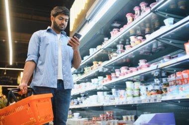 Akıllı telefon kullanan ve markette süt ürünlerini seçerken alışveriş sepeti taşıyan odaklanmış Hintli bir adam.