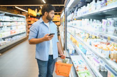 Genç Hintli bir adam akıllı telefon kullanıyor ve marketten alışveriş sepeti tutuyor.