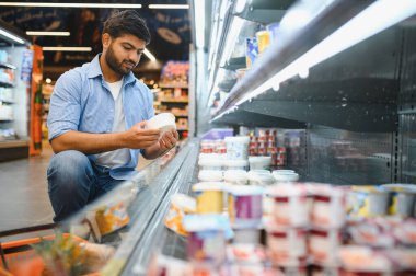 Genç Hintli bir adam markette alışveriş yaparken bir süt ürünü etiketi üzerinde malzemeleri inceliyor, beslenme ve kaliteye odaklanıyor.