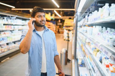 Genç Hintli bir adam alışveriş arabasını itiyor ve süpermarkette yiyecek seçerken telefonla konuşuyor.