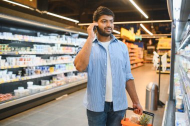 Genç Hintli bir adam alışveriş sepeti taşırken bir telefon görüşmesi yapıyor, hareketli bir marketin koridorlarında geziniyor.