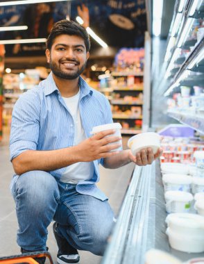 Genç Hintli bir adam süpermarketin mandırasında çömelmiş iki yoğurt konteynırını karşılaştırıyor.