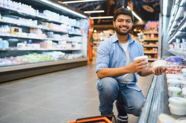 Gülümseyen bir müşteri marketteki soğutulmuş rafın yanında çömelmiş iki yoğurt konteynırını karşılaştırıyor.