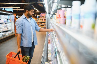 Buzdolabından süt ürünlerini seçen konsantre genç Hintli adam süpermarkette ananas dolu alışveriş sepeti tutuyor.