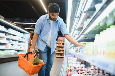 Hintli müşteri, süpermarketten alışveriş yaparken, soğutulmuş raflardan bir ürün seçiyor, ananaslı bir alışveriş sepeti tutuyor,