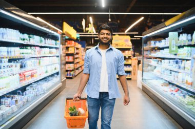 Bir marketin koridorunda yürürken elinde alışveriş sepeti taşıyan genç Hintli bir adam.