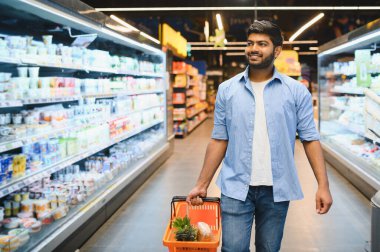 Genç Hintli bir adam süpermarket koridorlarında yürürken bir alışveriş sepeti taşıyor, içinde bakkaliye bulunan soğutulmuş raflara bakıyor.