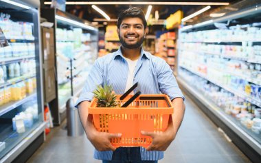 İçinde ananas olan turuncu bir alışveriş sepeti tutan genç Hintli adam, bakkal koridorunda gülümsüyor.