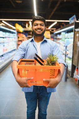 Ananaslı bir alışveriş sepeti taşıyan genç bir Hintli süpermarkette alışveriş yaparken gülümsüyor.
