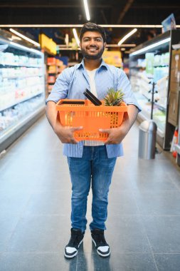 Genç Hintli bir adam içinde ananas olan bir alışveriş sepeti taşıyor, bakkal koridorunda gülümsüyor.