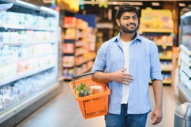 Genç Hintli bir adam alışveriş sepeti taşıyor, hareketli bir süpermarketin parlak ve modern koridorlarında geziniyor.