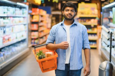Parlak ve düzenli bir süpermarkette alışveriş yaparken alışveriş yaparken alışveriş sepetini taşıyan genç Hintli bir adam perakende deneyiminin tadını çıkarıyor.