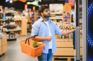 Genç Hintli bir adam, açık ve düzenli bir market koridorunda buzdolabının kapısını açarken elinde alışveriş sepeti taşıyor.