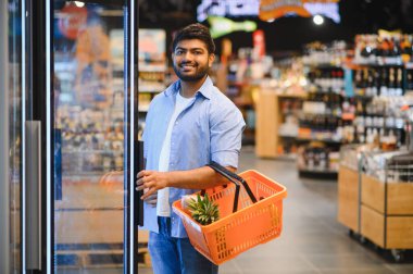 Genç Hintli bir adam elinde bir alışveriş sepeti ve açık buzdolabı kapısı parlak, organize bir market koridorunda