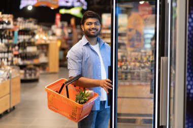 Genç Hintli bir adam markette buzdolabının kapısını açıyor, içinde ananas olan bir alışveriş sepeti tutuyor.