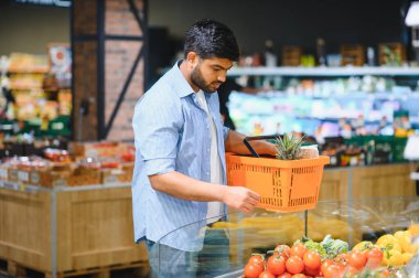 Genç Hintli bir adam süpermarketten taze sebze seçiyor, içinde ananas olan turuncu bir alışveriş sepeti tutuyor.
