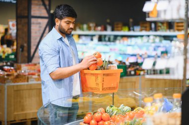Odaklanmış müşteri taze domates seçiyor, süpermarkette alışveriş sepeti tutuyor, sağlıklı yiyecek seçimleri yapıyor.