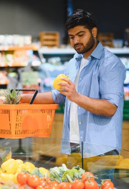 Genç Hintli bir adam markette alışveriş sepeti tutarken sarı dolmalık biber seçiyor.