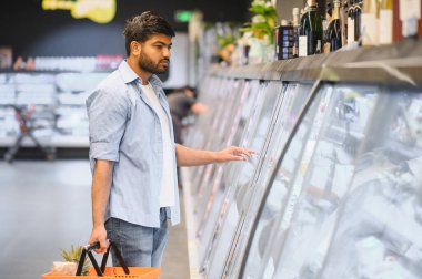 Müşteri marketin dondurulmuş bölümünden sebze seçiyor, elinde ananas dolu alışveriş sepeti tutuyor.