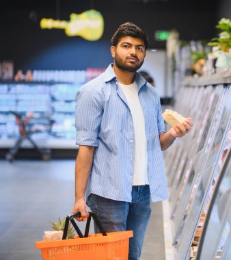 Müşteri alışveriş sepetini tutuyor ve marketin soğutulmuş bölümünden peynir seçiyor.