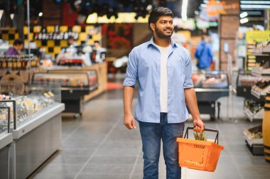Bir süpermarket koridorunda yürürken elinde alışveriş sepeti taşıyan genç Hintli bir adam taze ürünler seçiyor.