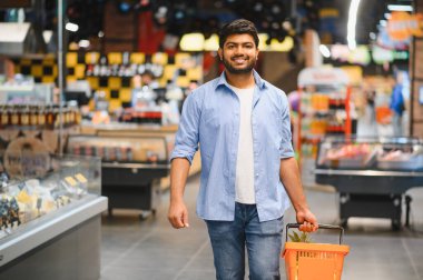 Market koridorlarında yürürken alışveriş deneyiminin tadını çıkaran bir alışveriş sepeti taşıyan genç Hintli adam.