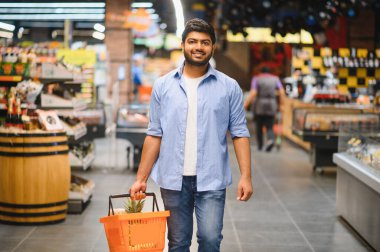 Market koridorunda yürürken alışveriş sepetini taşıyan gülümseyen Hintli adam alışveriş deneyiminin tadını çıkarıyor.