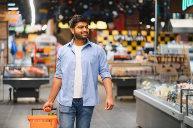 Genç Hintli adam elinde alışveriş sepetiyle süpermarkette yürüyor, gülümsüyor ve etrafa bakıyor.