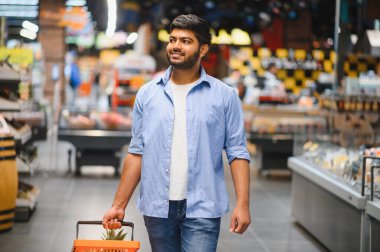 Bir süpermarket koridorunda yürürken elinde alışveriş sepeti taşıyan genç bir Hintli.