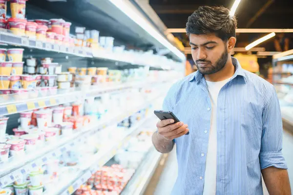 Market fiyatlarını kontrol etmek için akıllı telefon kullanan ya da soğutulmuş rafların yanında yürüyen süpermarketteki ürünleri karşılaştıran Hintli bir müşteri.