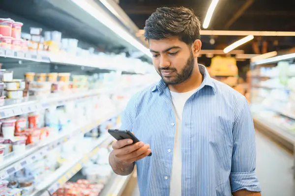 Markette alışveriş yaparken akıllı telefon kullanan, alışveriş listesini kontrol eden ya da fiyatları karşılaştıran Hintli bir adam.