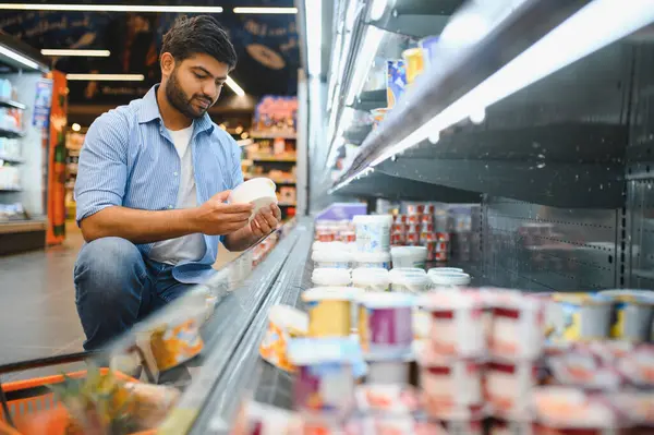Genç Hintli bir adam markette alışveriş yaparken bir süt ürünü etiketi üzerinde malzemeleri inceliyor, beslenme ve kaliteye odaklanıyor.