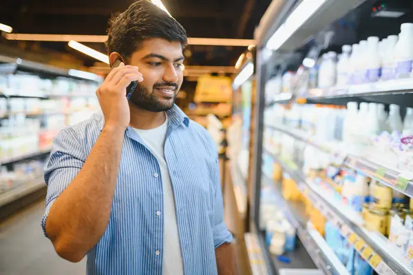 Genç Hintli adam süpermarketten alışveriş yaparken cep telefonuyla konuşuyor, süt ürünlerini seçiyor.
