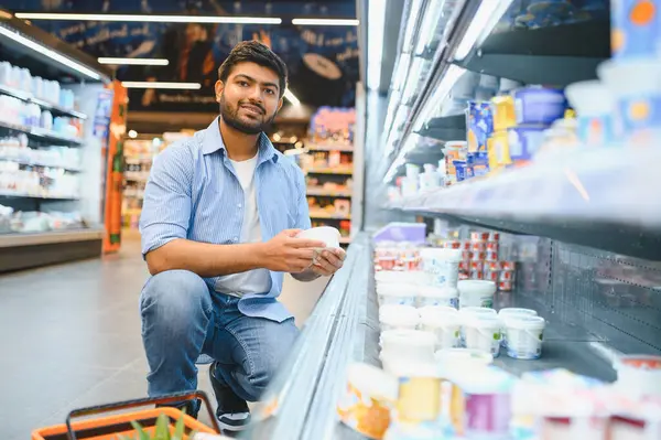 Genç adam, çeşitli seçeneklerle çevrili bir süpermarketin mandıra koridorunda alışveriş yaparken çömelmiş yoğurt konteynırını tutuyor.